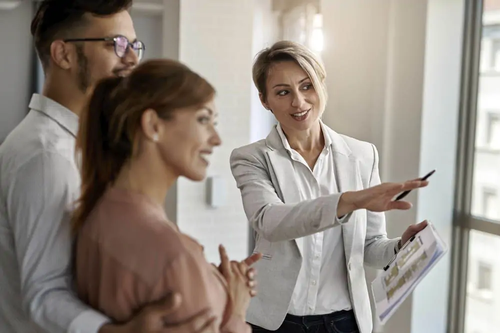 Um homem abraçando uma mulher à esquerda e a direita uma mulher com um documento e canetas na mão explicando algo