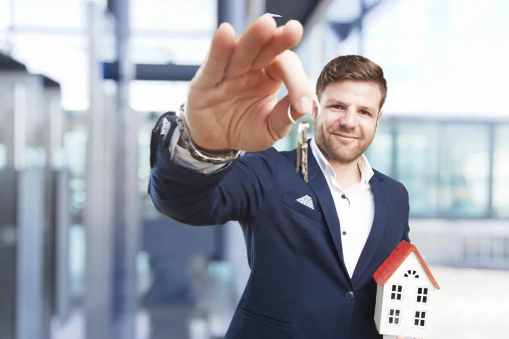 Homem segurando um par de chaves com o braço esquerdo levantado e uma mini casa ao lado direito.