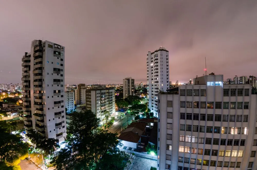 Visão noturna de edifícios residenciais na cidade de São Paulo, iluminados com luzes azuis, verdes e brancas, sob céu nublado.