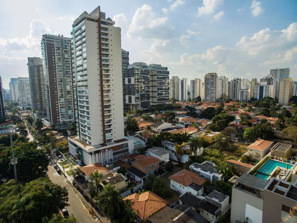 Vista de uma cidade com prédios altos modernos e casas menores residenciais, com piscina e muitas árvores, sob céu parcialmente nublado.