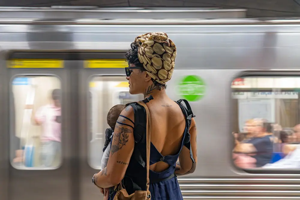 Mulher elegante, usando turbante estampado e óculos escuros, espera no metrô movimentado, segurando o filho no colo em meio à agitação urbana.